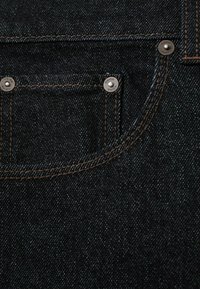 Tela de mezclilla oscura con costuras marrones en contraste, un pequeño bolsillo delantero y detalles de remaches plateados. Textura suave con un ligero brillo.