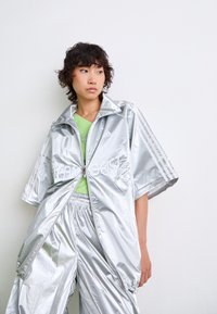 Persoon in een glanzend zilveren Adidas trainingspak met losse korte mouwen en broek, met een limoengroene top eronder, staand tegen een witte muur.