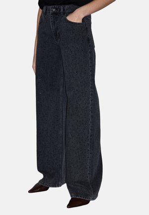 Person trägt hoch taillierte, weit geschnittene schwarze Jeanshosen mit dezentem Pünktchenmuster und braune spitze Schuhe, eine Hand in der Tasche, vor einfarbigem Hintergrund.