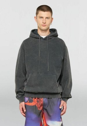 Giovane uomo che indossa una felpa nera sbiadita con cappuccio e pantaloncini colorati viola e rossi con motivi, in piedi davanti a uno sfondo grigio semplice.