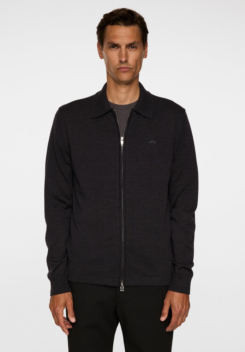 Uomo che indossa un maglione grigio scuro con zip e colletto sopra una camicia grigia, in piedi contro uno sfondo bianco semplice.