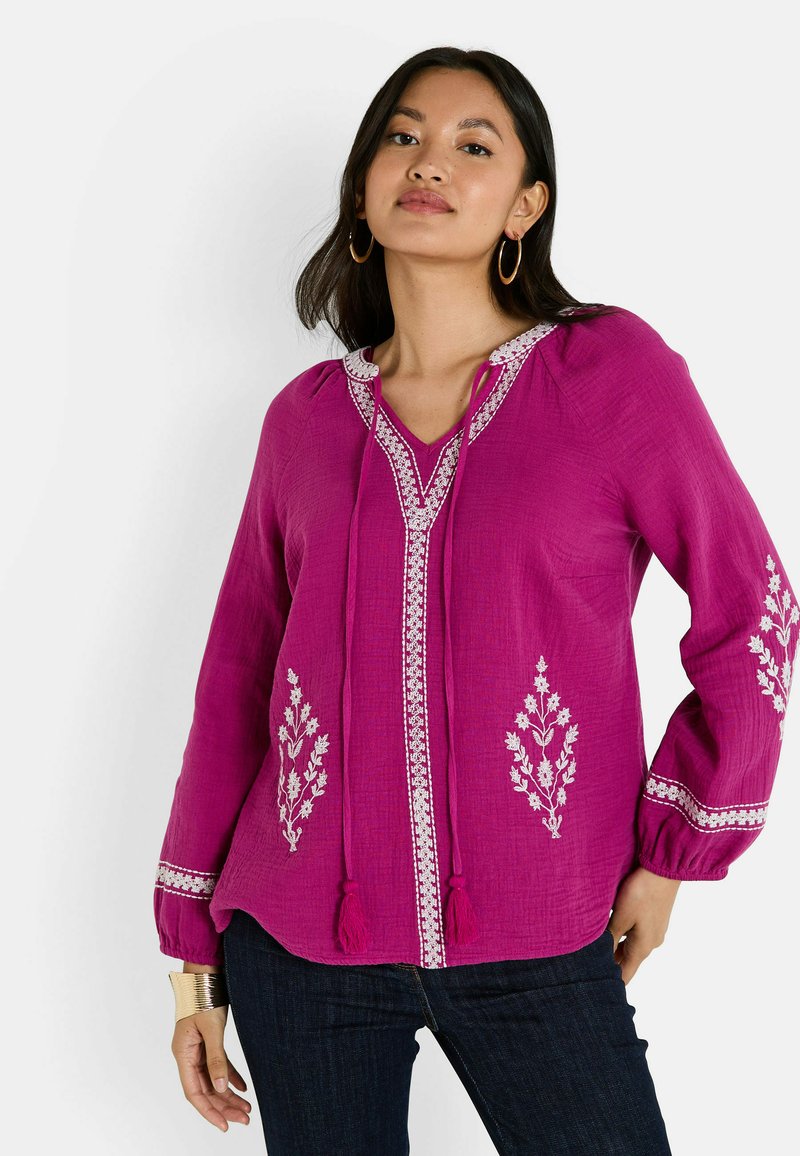 Blouse fuchsia en tissu léger, avec broderie florale, décolleté en V et liens à pompons. Manches longues avec poignets élastiques.
