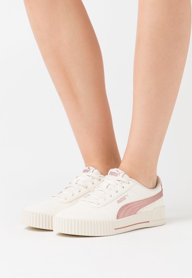 Baskets blanches Puma avec un détail bande rose portées aux pieds sur un fond blanc uni.