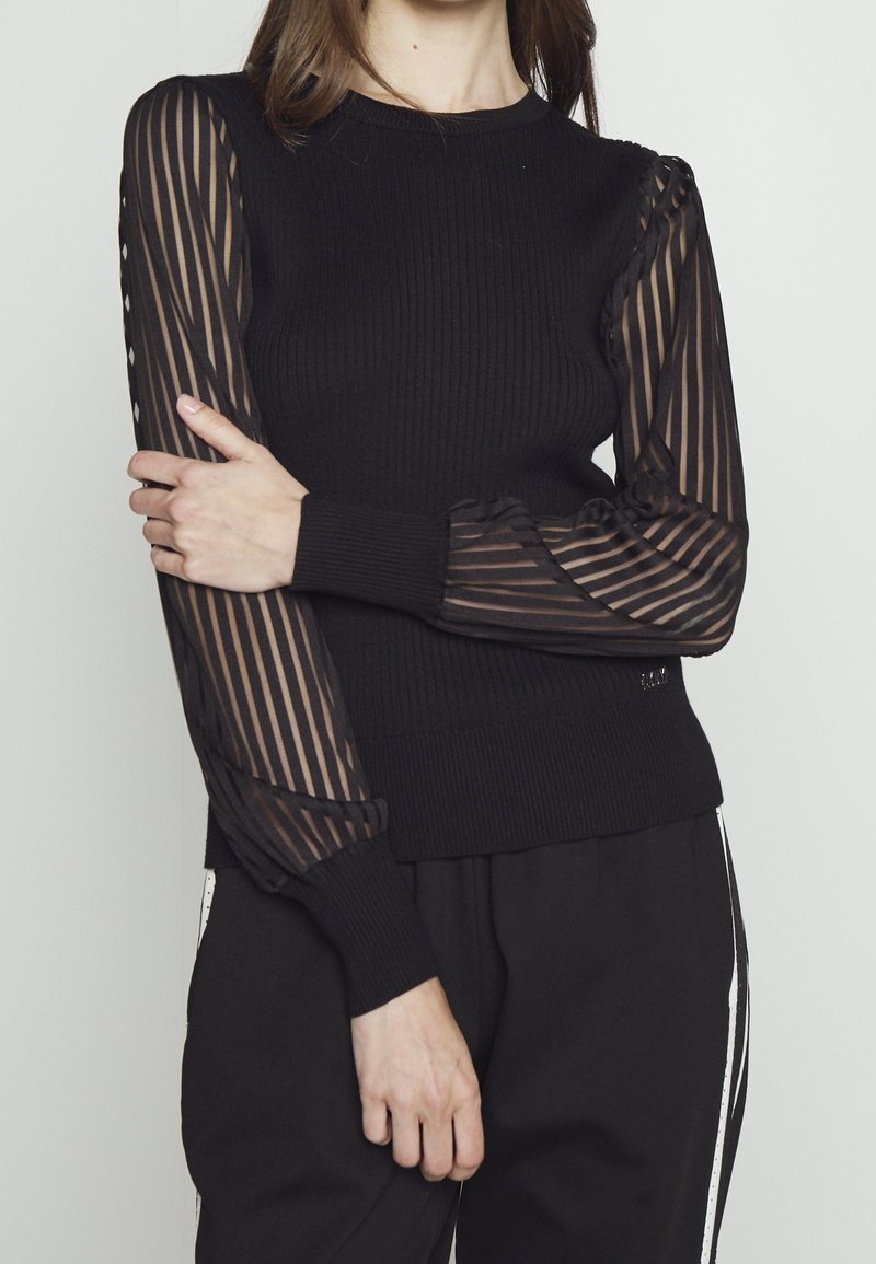 Mujer con suéter negro acanalado de mangas largas transparentes con rayas, combinada con pantalones negros con rayas laterales blancas, de pie frente a un fondo liso.