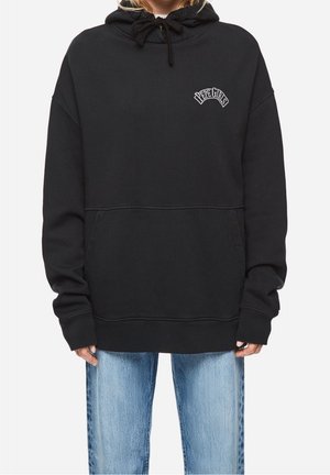 Personne portant un sweat à capuche noir oversize avec le texte "PEPE GIRLS" sur la poitrine et un jean bleu clair sur un fond uni.