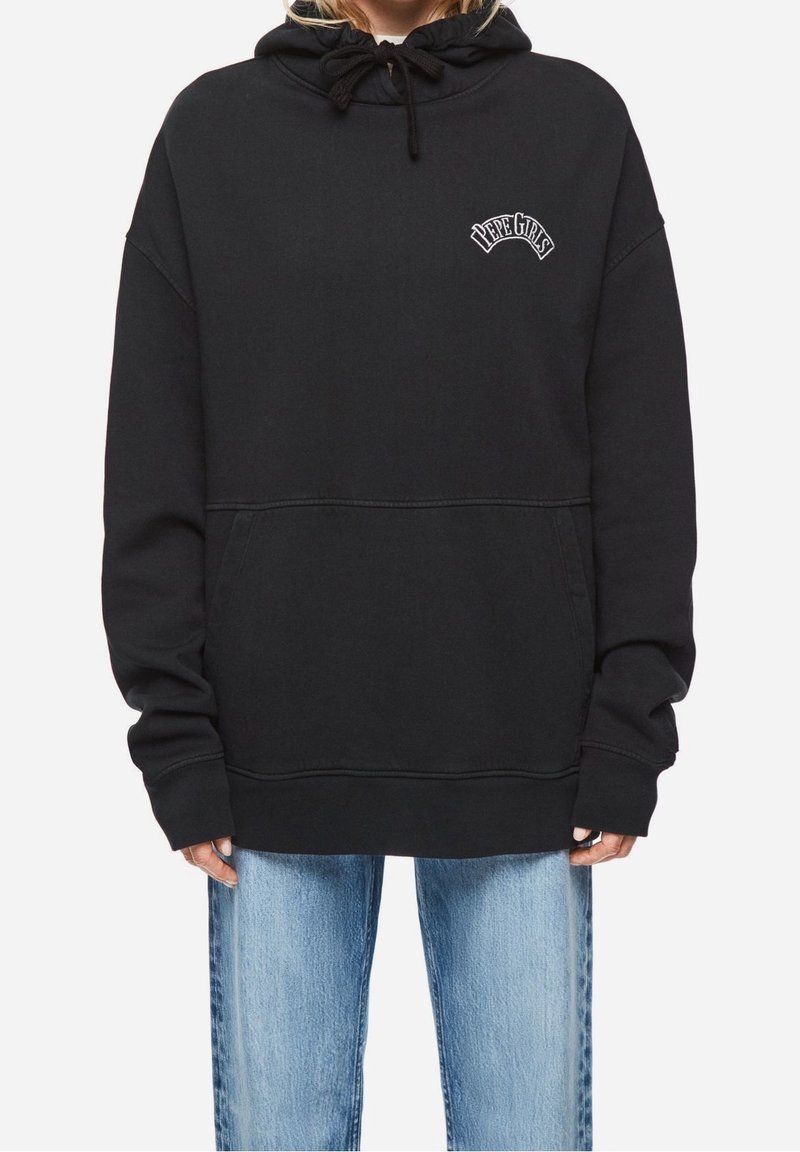 Personne portant un sweat à capuche noir oversize avec le texte "PEPE GIRLS" sur la poitrine et un jean bleu clair sur un fond uni.