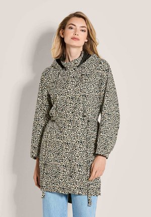 Vrouw draagt een lange luipaardprint capuchonjas met trekkoorden en blauwe jeans, staand tegen een eenvoudige neutrale achtergrond.