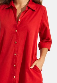 Chemise rouge à manches 3/4, avec col et poches. Confectionnée en tissu léger, elle présente des boutons dorés avec un motif floral.