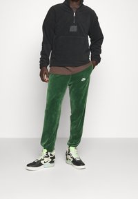 Persona che indossa un hoodie nero a metà zip, una maglietta marrone sotto, pantaloni sportivi verdi Nike e sneakers alte bianco e nere con dettagli verdi.