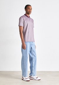 Polo shirt viola con colletto e dettagli ricamati, abbinato a jeans a gamba dritta blu chiaro e sneakers bianche con accenti rossi.