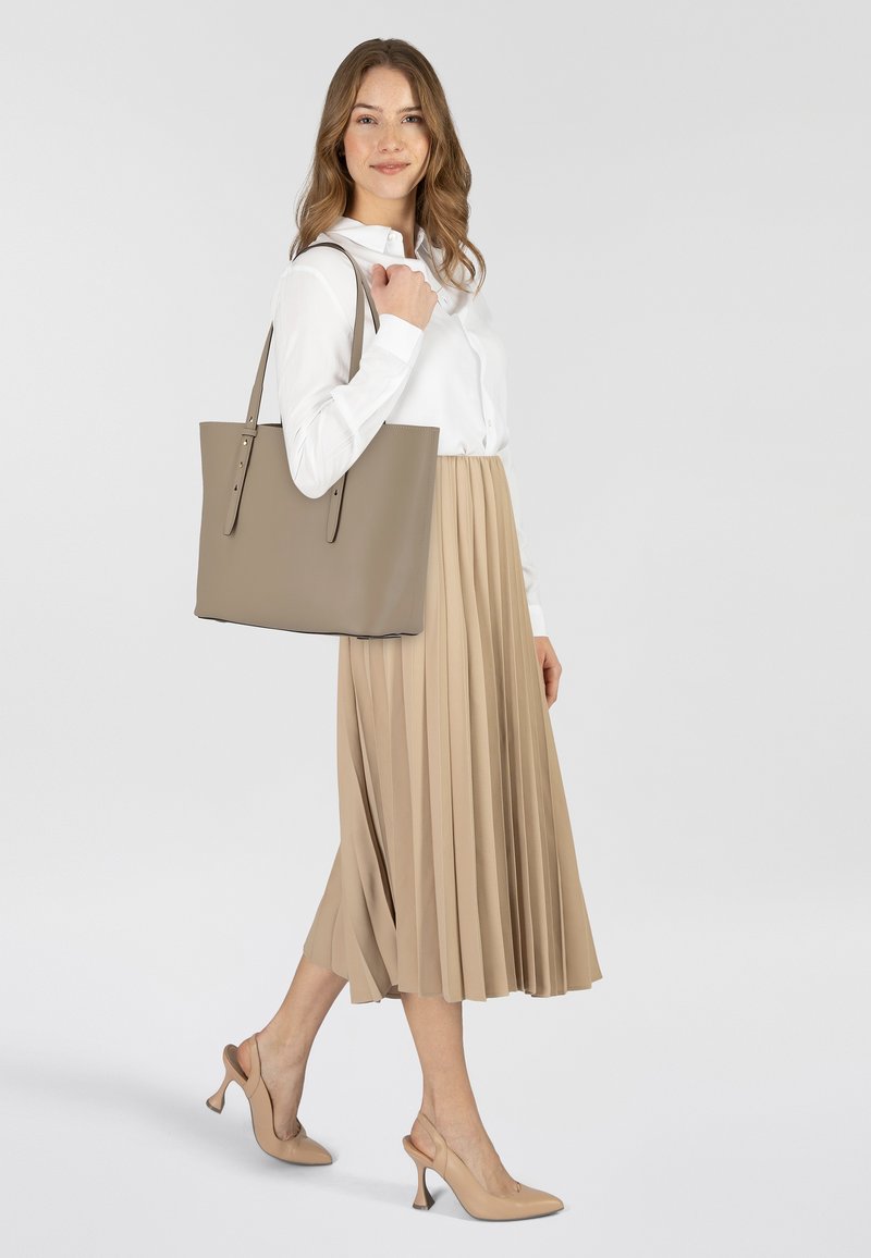Beige leren totebag met twee schouderriemen, minimalistisch ontwerp, gladde textuur, gecombineerd met een wit overhemd en een geplooide beige rok.