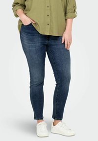 Vaqueros ajustados de color azul oscuro con una textura de denim suave. Combinados con una camisa verde de botones y zapatillas deportivas blancas. Detalles visibles de los bolsillos laterales.