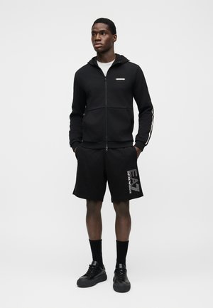 Μαύρο φούτερ με φερμουάρ και κουκούλα, με λογότυπο στο στήθος. Συνδυάζεται με μαύρα σορτς που φέρουν λευκό γραφικό. Μαύρα αθλητικά παπούτσια και κάλτσες μέχρι τον αστράγαλο ολοκληρώνουν το σύνολο.