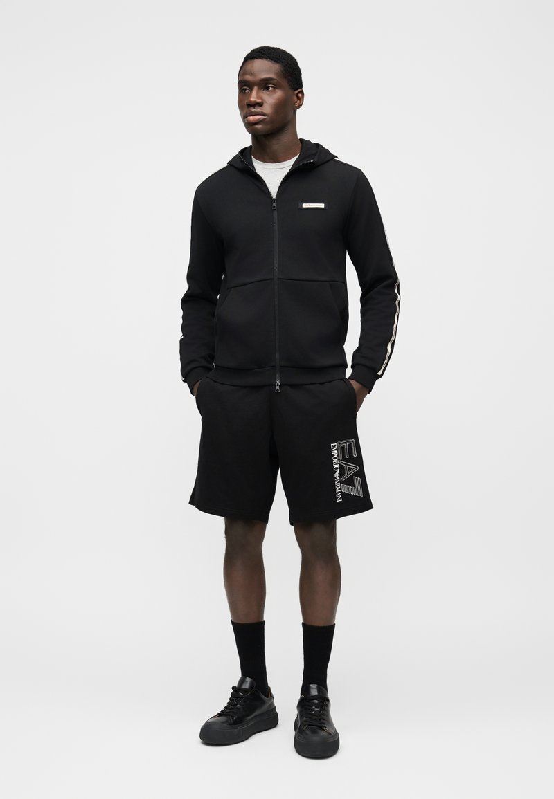 Μαύρο φούτερ με φερμουάρ και κουκούλα, με λογότυπο στο στήθος. Συνδυάζεται με μαύρα σορτς που φέρουν λευκό γραφικό. Μαύρα αθλητικά παπούτσια και κάλτσες μέχρι τον αστράγαλο ολοκληρώνουν το σύνολο.