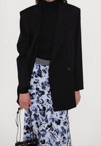 Černý oversized blejzr se strukturovanými rameny, nošený přes černý top, doplněný modro-bílou vzorovanou sukní a malou černou kabelkou.