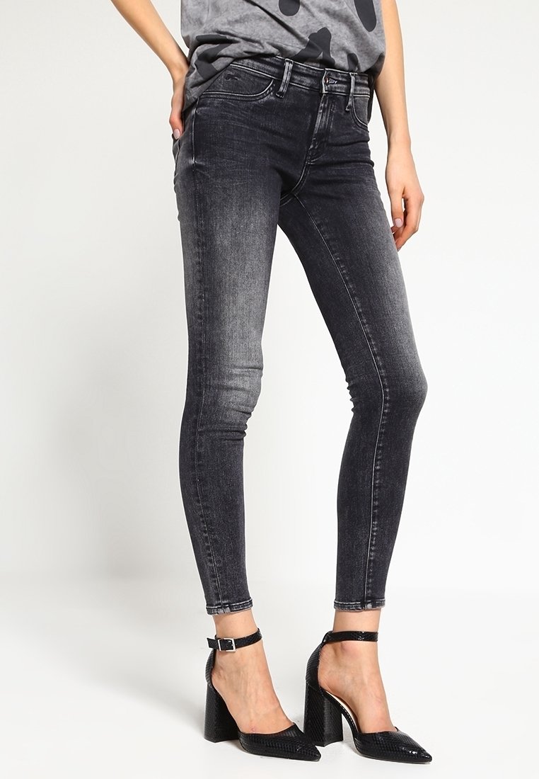 Svarta smala jeans med en blekt design, framfickor och bälteshällor, tillsammans med svarta klackskor med ankelremmar på en neutral bakgrund.