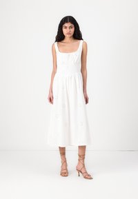 Abito midi bianco con scollatura quadrata, motivi floreali ricamati e vita aderente. Il modello indossa sandali beige con cinturini.