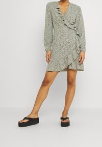 Blumendruck Wickelkleid in Hellgrün mit Rüschendetail, langen Ärmeln und schwingendem Saum, kombiniert mit schwarzen Plateau-Sandalen.