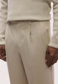 Beige bukser med skreddersydd passform, med to frontfolder og sidelommer, laget av glatt stoff, kombinert med en kremfarget ribbestrikket genser.