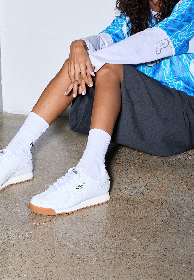 Personne assise sur un sol en béton portant des baskets blanches Puma, des chaussettes blanches montantes, un short foncé et un haut à manches longues bleu à motifs.