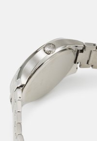 Reloj de acero inoxidable plateado con caja redonda, textura cepillada y un detalle de corona prominente. Presenta un diseño de pulsera articulada.