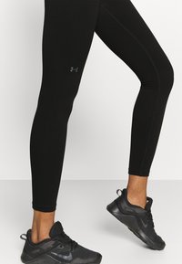 Schwarze Sportleggings mit glatter Textur, ausgestattet mit einem kleinen grünen Emblem am Oberschenkel und einem figurbetonten Design. Kombiniert mit schwarzen Sneakers.