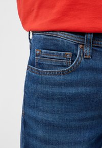 Blaues Jeans-Taschen aus Denim mit orangefarbenen Nähten, getragen zu einem in die Hose gesteckten roten Hemd.