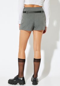 Graue, maßgeschneiderte Shorts mit einer taillierten Passform und einem schwarzen Akzentgürtel. Kombiniert mit knielangen schwarzen Socken und klobigen schwarzen Schuhen.