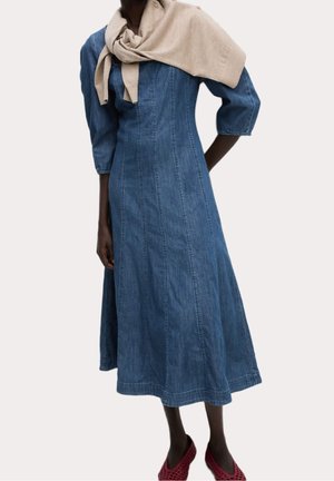 Kvinde iført en blå denim midi-kjole med trekvartærmer, en beige sweater draperet over skuldrene og røde flettede sko.