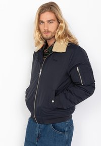 Chaqueta bomber de color navy con un cuello de piel sintética beige, cierre en la parte delantera y puños de canalé. Cuenta con un bolsillo con cremallera en la manga y un ajuste relajado.