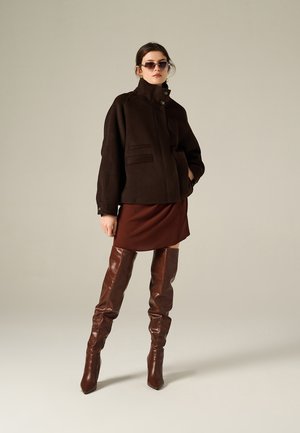 Manteau en laine brun surdimensionné avec col haut, fermeture à boutons sur le devant et deux poches latérales ; assorti à une robe mini brune et des bottes en cuir montantes.