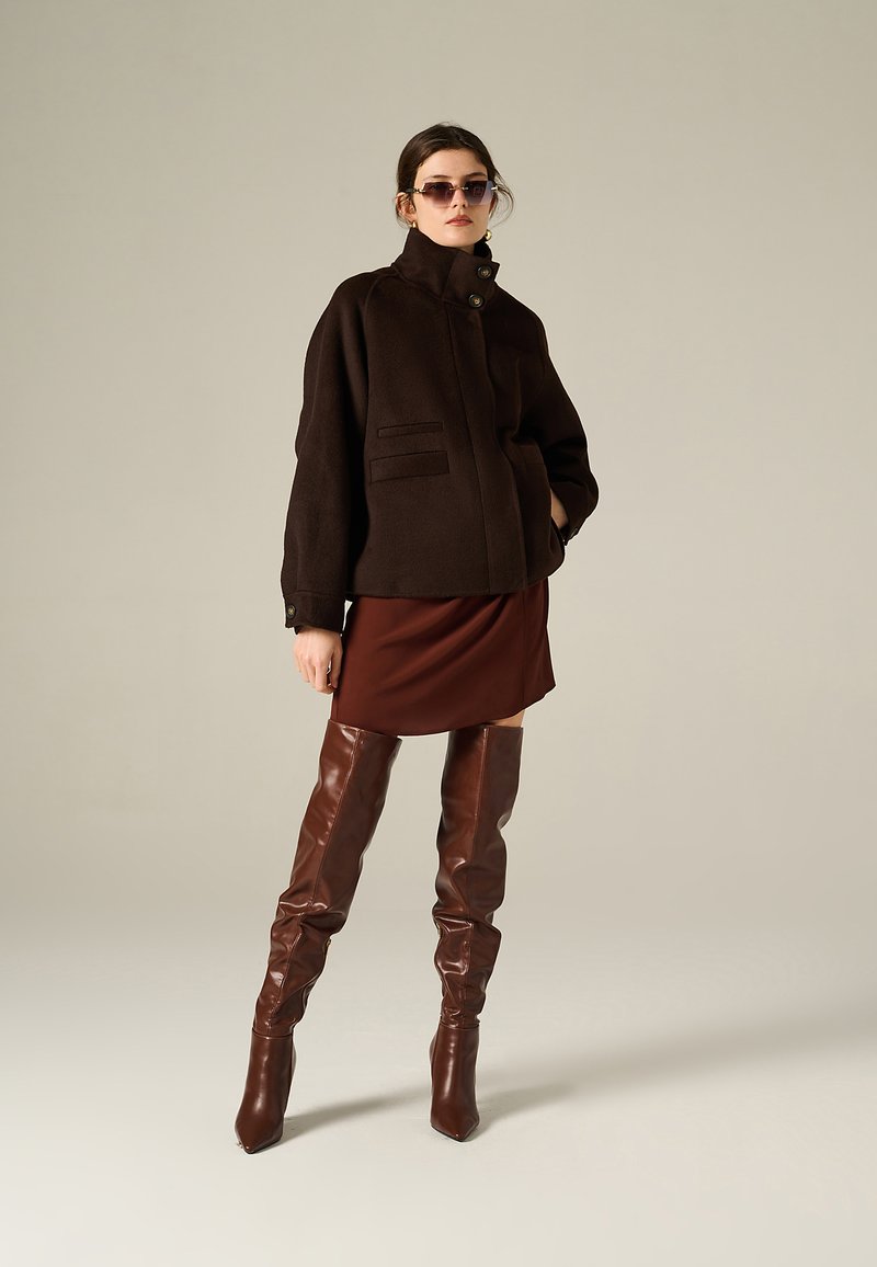 Cappotto oversized marrone in lana con colletto alto, chiusura frontale con bottoni e due tasche laterali; abbinato a un mini abito marrone e stivali in pelle alti fino alla coscia.