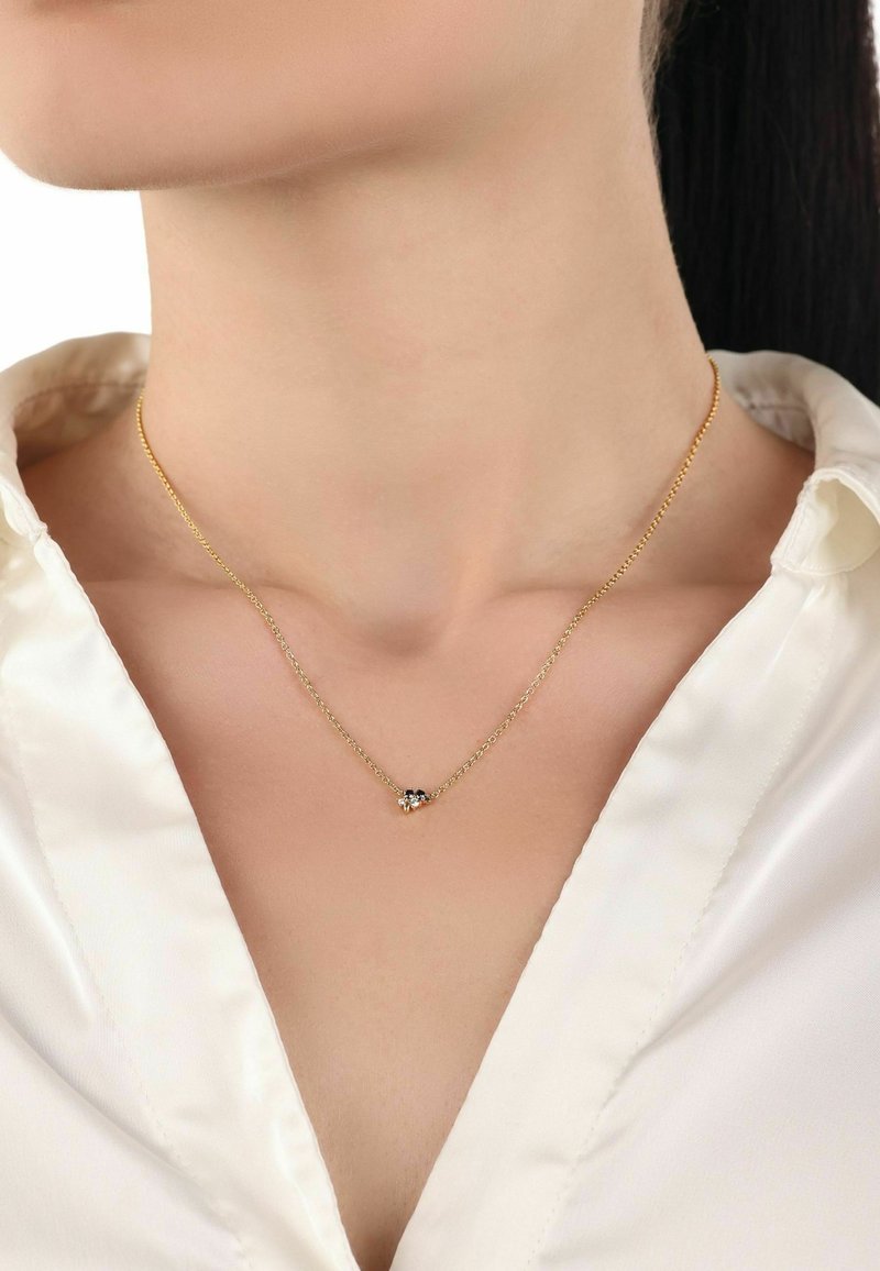 Złoty łańcuszek z małym kryształowym wisiorkiem, o delikatnym wzornictwie, gładkiej fakturze i regulowanej długości, noszony na białej bluzce.