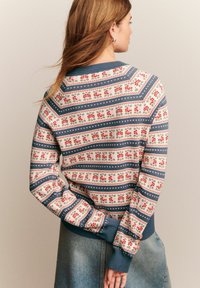 Vrouw met lang haar die een gebreide trui met rode en blauwe bloem- en geometrische ontwerpen draagt, staand met haar handen achter haar rug.