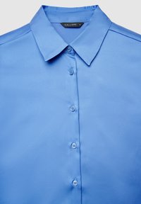 Light blue, collared button-up shirt with five buttons down the front and a black label reading "CALLIOPE S" inside the collar.