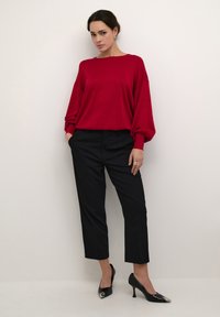 Crvena pletena majica s slobodnim rukavima, uparena s crnim krojenim hlačama. Crne cipele s oštrim vrhom dodaju elegantan detalj. Jednostavna pozadina.