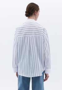 Camisa blanca de manga larga con rayas verticales azul marino, puños abotonados y diseño de yugo en la parte posterior, hecha de material ligero.