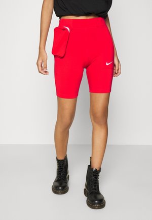 Pantaloncini da bici Nike rossi con tasca laterale e borsello annesso, indossati con stivali combat neri e top corto nero su sfondo grigio chiaro.