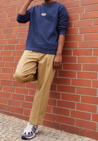 Marinblå sweatshirt med "Levi's"-logotyp, beige plisserade byxor och gråsvarta sneakers med vita detaljer, mot en tegelvägg.