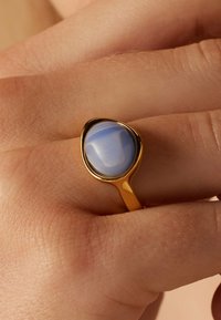 Gouden ring met een druppelvormige lichtblauwe steen. Glad, gepolijst oppervlak met een glanzende afwerking. Draagt aan een hand met een natuurlijke huidskleur.