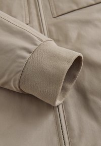 Beige jakkeærme tæt på, med ribbet manchet, glat tekstur og en lodret lynlås. Subtile syninger fremhæver designet.