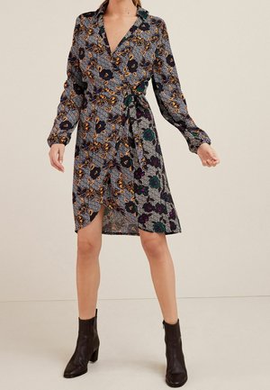 Femme portant une robe longue à manches longues de style portefeuille à motifs floraux, avec des bottines noires, debout devant un fond beige uni.