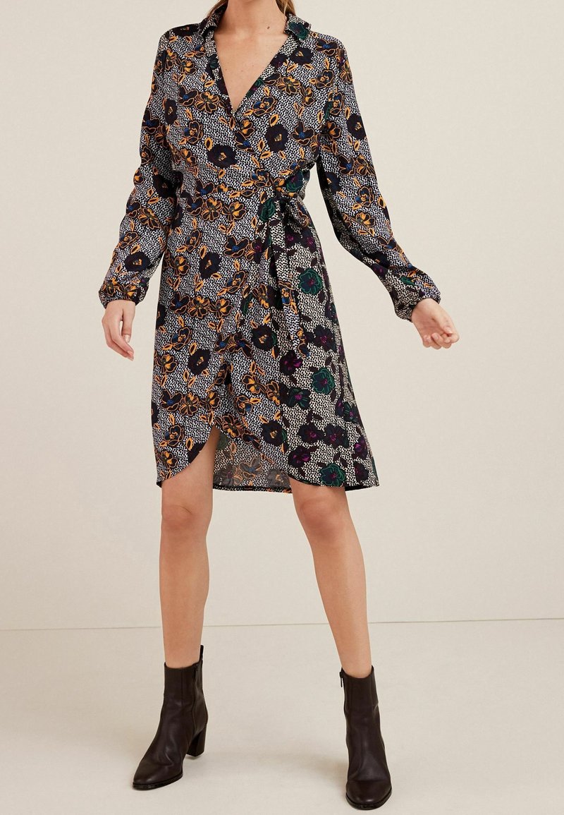 Femme portant une robe longue à manches longues de style portefeuille à motifs floraux, avec des bottines noires, debout devant un fond beige uni.