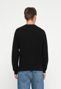 Egy férfi háttal áll, fekete bordázott kötött pulóverben és kék farmernadrágban, egy sima fehér háttér előtt.
