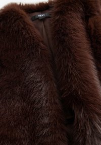 Gros plan sur le col d'un manteau en fausse fourrure brun foncé avec une texture douce et une étiquette noire indiquant le nom de la marque "NEXT."