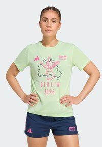 Vaalean vihreä urheilupaita, jossa on vaaleanpunainen ja musta graafinen printti; mukana teksti ja logo "Berliinin maraton 2025". Yhdistetty tummansinisiin shortseihin.