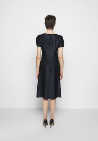 Žena stojící zády k fotoaparátu, oblečená v černých saténových šatech ke kolenům a černých vysokých podpatcích, na jednoduchém bílém pozadí.