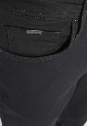 Krupan plan džepnog dijela crnih traperica s crnim metalnim oznakom marke na kojoj piše "ANTONY MORATO", zašivenom iznad otvora džepa.