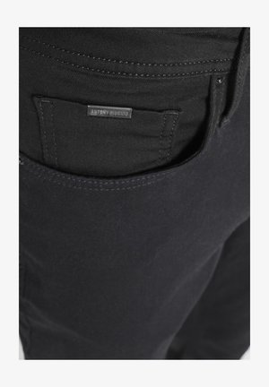 Close-up of black denim jeans pocket with black metal brand tag reading "ANTONY MORATO" sewn above the pocket opening.