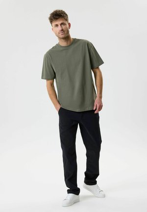 Moški, ki nosi majico kratkih rokavov olivno zelene barve, črne hlače in bele superge, stoji z rokami v žepih na enobarvnem ozadju.